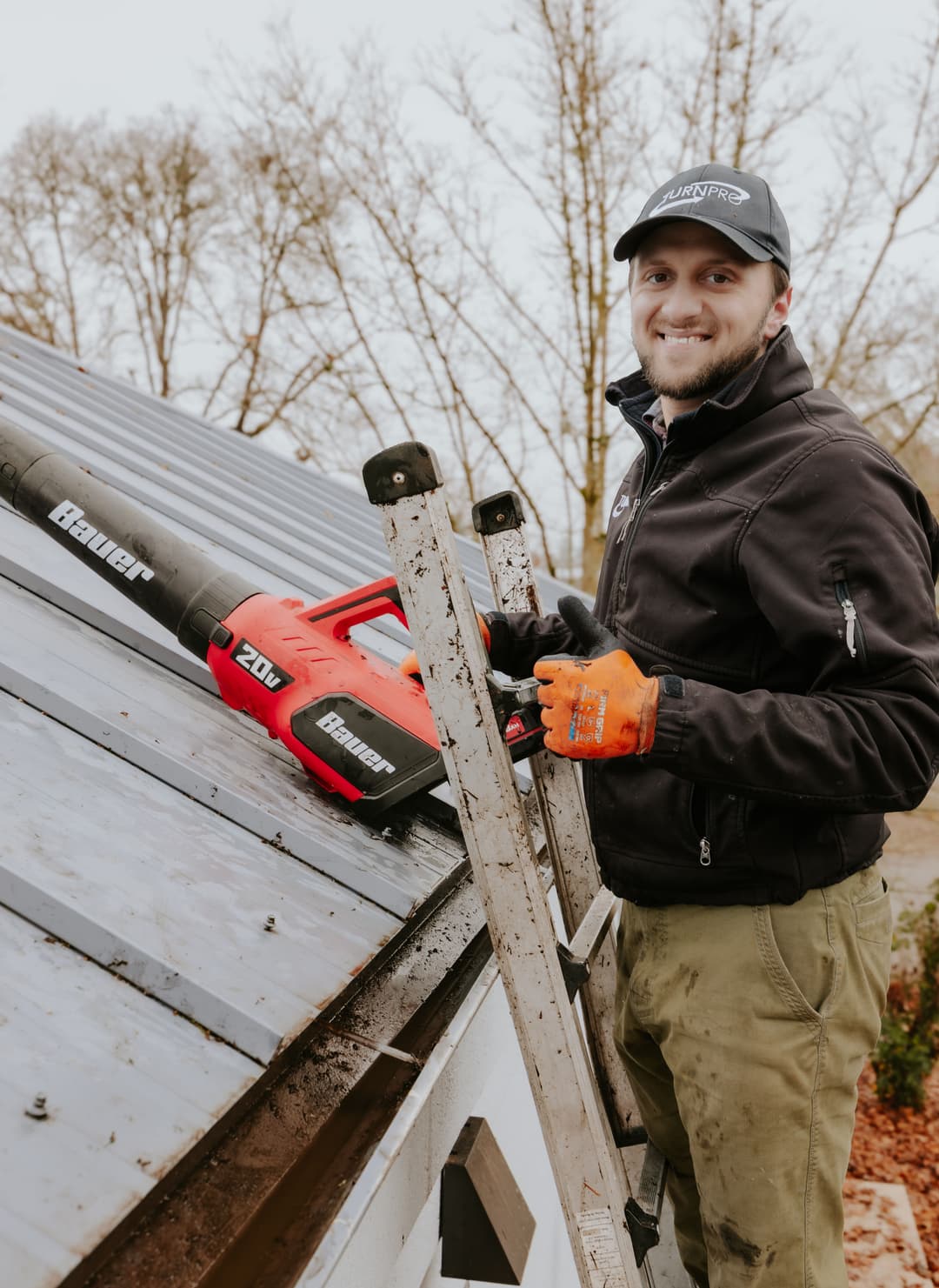 Man on a ladder cleaning roof gutters with a blower, wearing work gloves and a cap.
