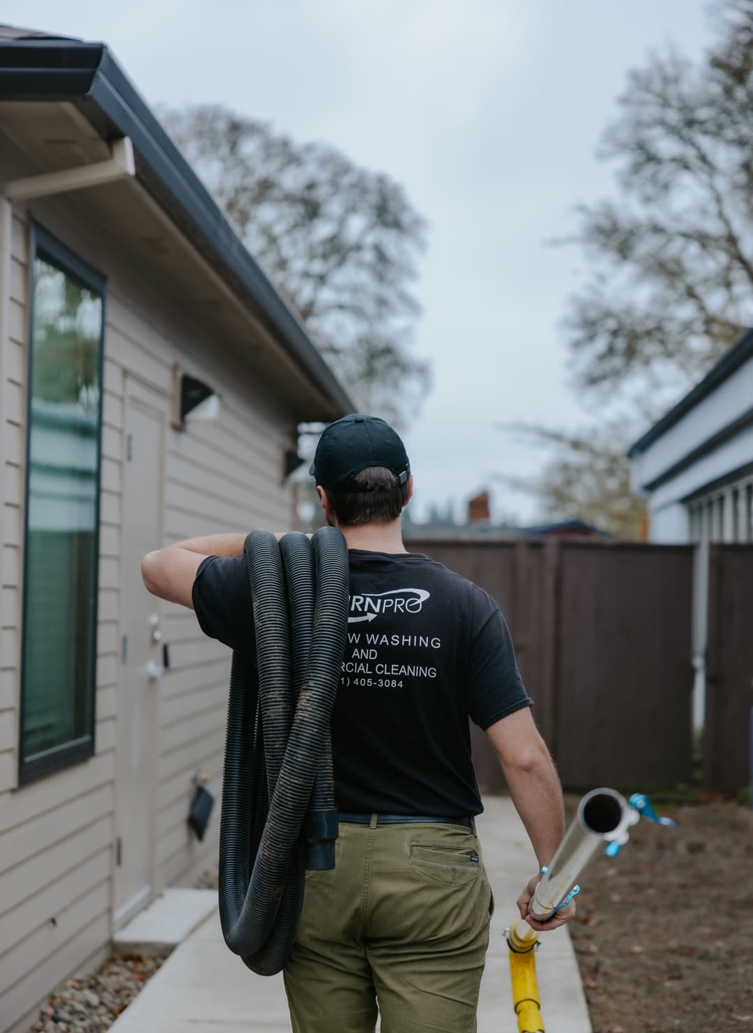 Person in a work uniform holding cleaning equipment walking beside a house.